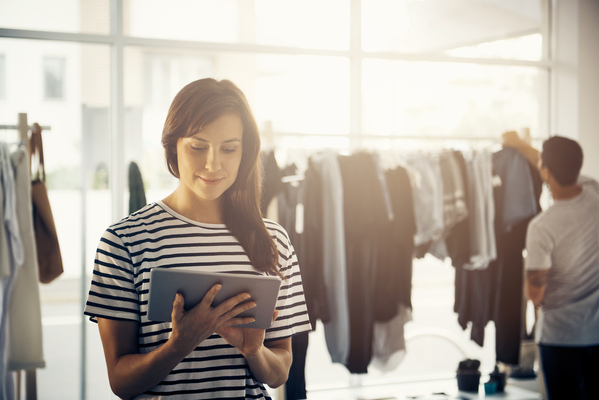 Emprendedora frente un rack de ropa con una tablet en la mano, mientras un cliente va la mercancía