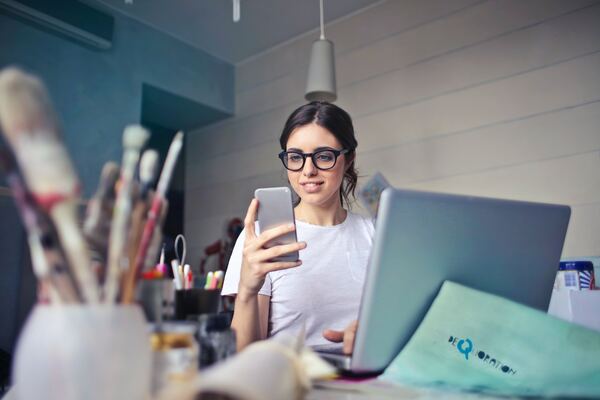 Community manager con el celular en la mano, la laptop sobre el escritorio y sonriendo a la cámara